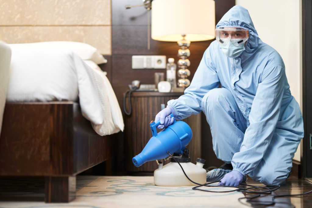 Worker in biohazard suit and mask disinfecting bedroom